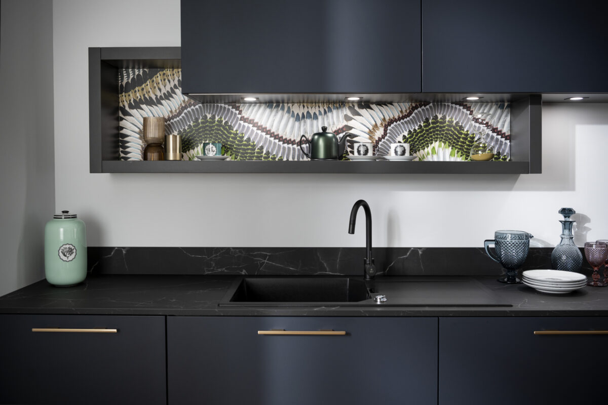 Das Federmotiv wird nochmal im Bereich der offenen Regale an Regalrückwand eingesetzt. Die marmorierte schwarze Arbeitsplatte ergänzt den eleganten Look der Designküche.