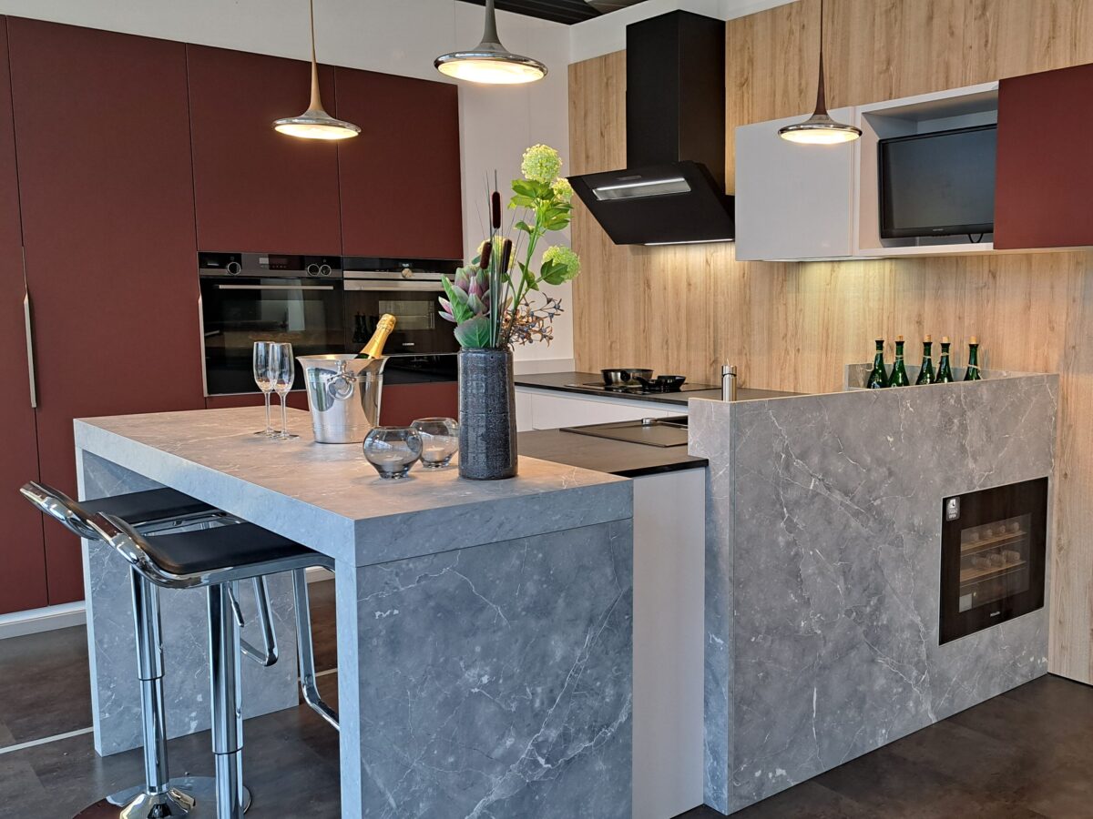 Moderne Ausstellungsküche mit einem angesetzen Barelement aus grauer Marmoroptik, zwei Barhockern, Weingläsern, einer Flasche in einem Eiskübel und einer Blumenvase. Kastanienbraune und holzfarbene Schränke sorgen zusammen mit eleganten Geräten und Pendelleuchten für eine stilvolle Präsentation.