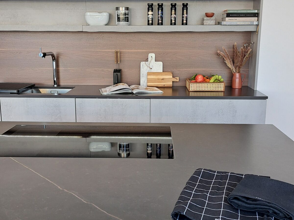 Moderne SCHMIDT Einbauküche mit brauner Keramikarbeitsplatte von Neolith, Spülbecken und offenen Regalen mit Schüsseln, Gewürzdosen, Büchern und Dekoration. Ein Geschirrtuch liegt gefaltet auf dem Tresen, frisches Gemüse in einem Korb neben einem Schneidebrett in der Nähe des Abflusses.