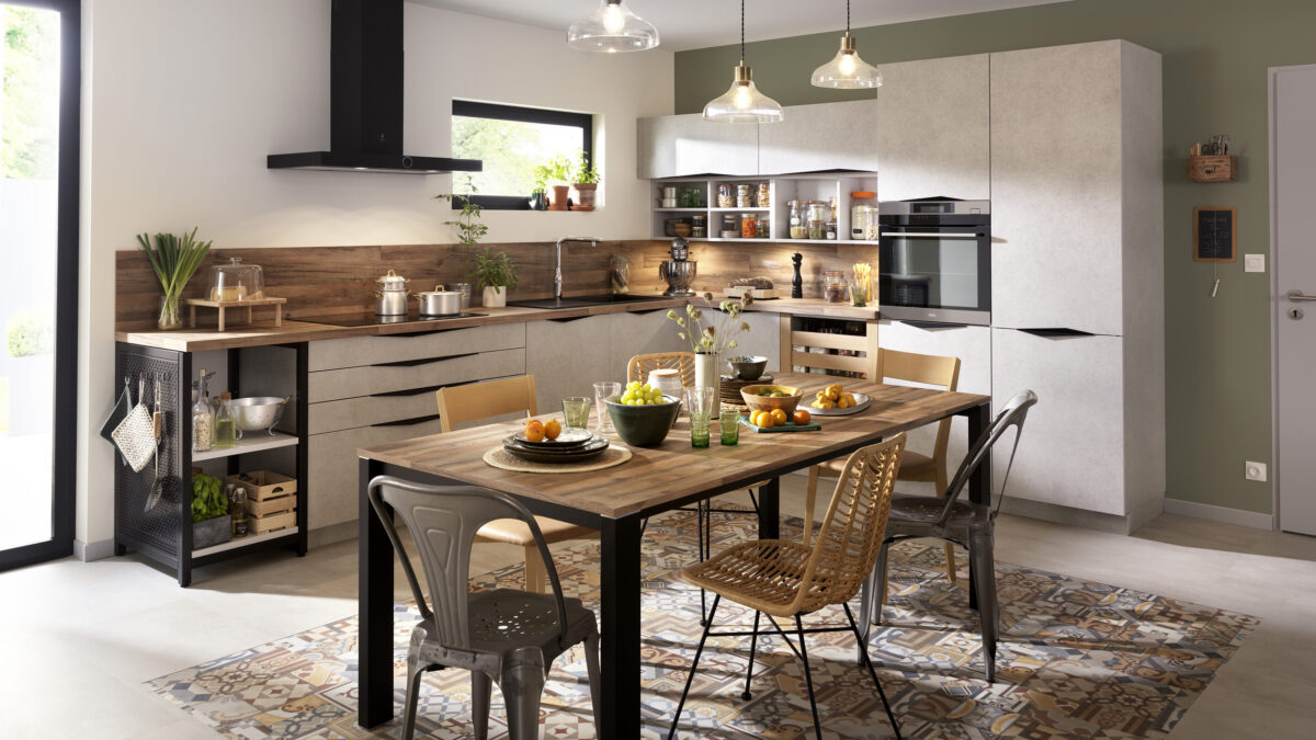 Klassische Küche mit hellen Schränken, Arbeitsplatten aus Holz und einem Esstisch, der für eine Mahlzeit gedeckt ist. Verschiedene Stühle umgeben den Tisch, und Hängelampen erhellen den Raum. Küchenutensilien und Pflanzen bringen Wärme in den Raum.