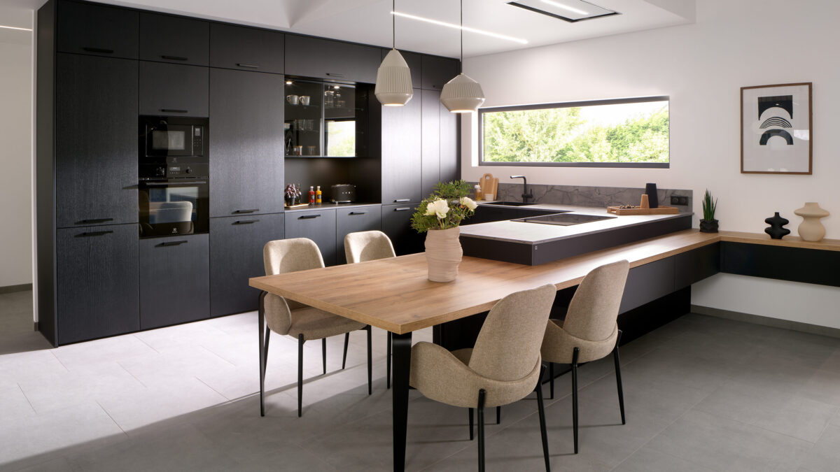 Moderne Designküche mit mattschwarzen Schränken, Einbaugeräten, einem Esstisch aus Holz mit vier beigen Stühlen, Pendelleuchten, einem großen Fenster und einer minimalistischen Einrichtung. Eine Vase mit Blumen steht auf dem Tisch.