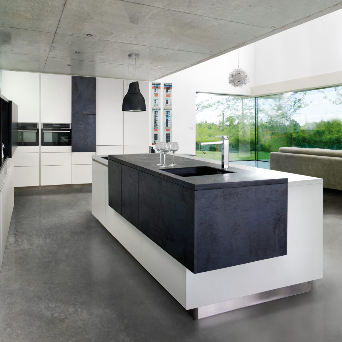 Moderne SCHMIDT Küche mit großer Kücheninsel, eingebautem Backofen, minimalistischen weißen Schränken, Hängeleuchten und bodentiefen Fenstern, die den Blick ins Grüne freigeben. Im Hintergrund ist ein Sofa zu sehen.