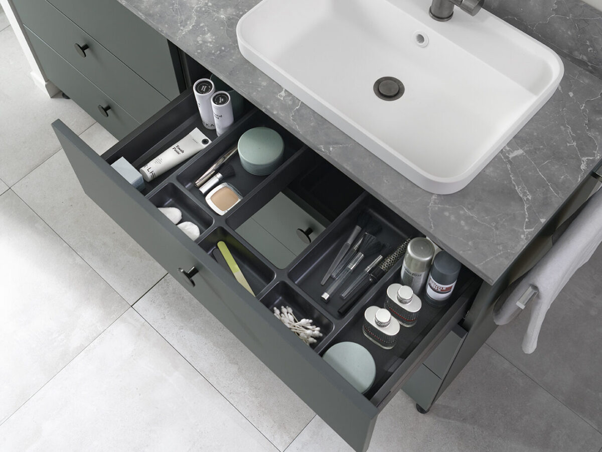 Eine offene Schublade unter einem Waschbecken in einem SCHMIDT Badezimmer. Darin befinden sich ordentlich organisiert verschiedene Toilettenartikel und Kosmetika wie Tiegeln, Tuben, Bürsten, Wattestäbchen und Flaschen, die alle in unterteilten Fächern angeordnet sind.