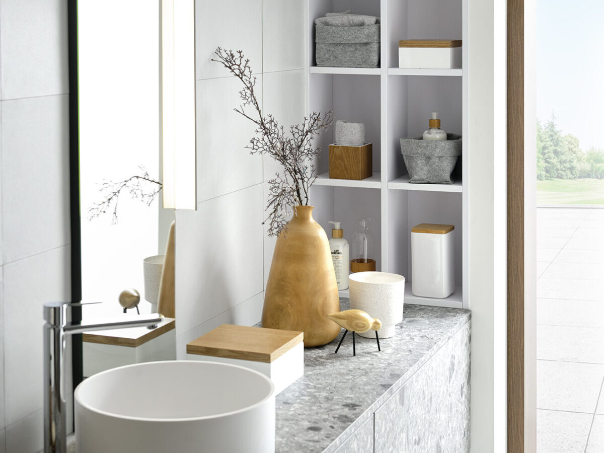 Modernes Badezimmer von SCHMIDT mit einem runden weißen Waschbecken, einem silbernen Wasserhahn und einer grauen Arbeitplatte in Terrazzo-Optik. Zu den dekorativen Elementen gehören eine große braune Vase mit Zweigen und eine kleine Vogelskulptur. Weiße Regale nehmen Handtücher und Behälter auf.