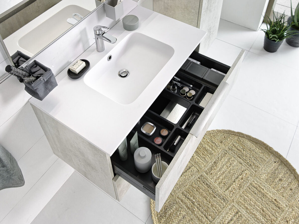Moderner SCHMIDT Badezimmer-Waschtisch mit einer herausziehbarer Schublade, in der Toilettenartikel, Make-up und Pflegeprodukte untergebracht sind. Ein weißes Waschbecken, Seife, Zahnbürstenhalter und Spiegel befinden sich auf dem Tresen; ein gewebter Teppich und Topfpflanzen sind auf dem weiß gefliesten Boden zu sehen.
