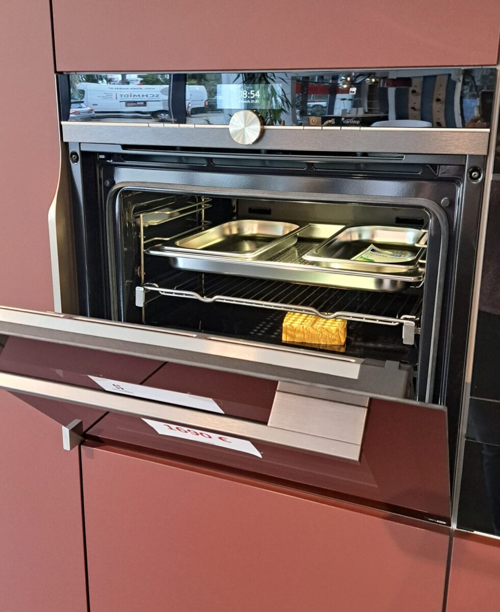 Ein Einbaubackofen mit teilweise geöffneter Tür, in der Backbleche und ein Laib Brot zu sehen sind. Der Backofen ist in einem kastanienbraunen Schrank mit modernem Design untergebracht.