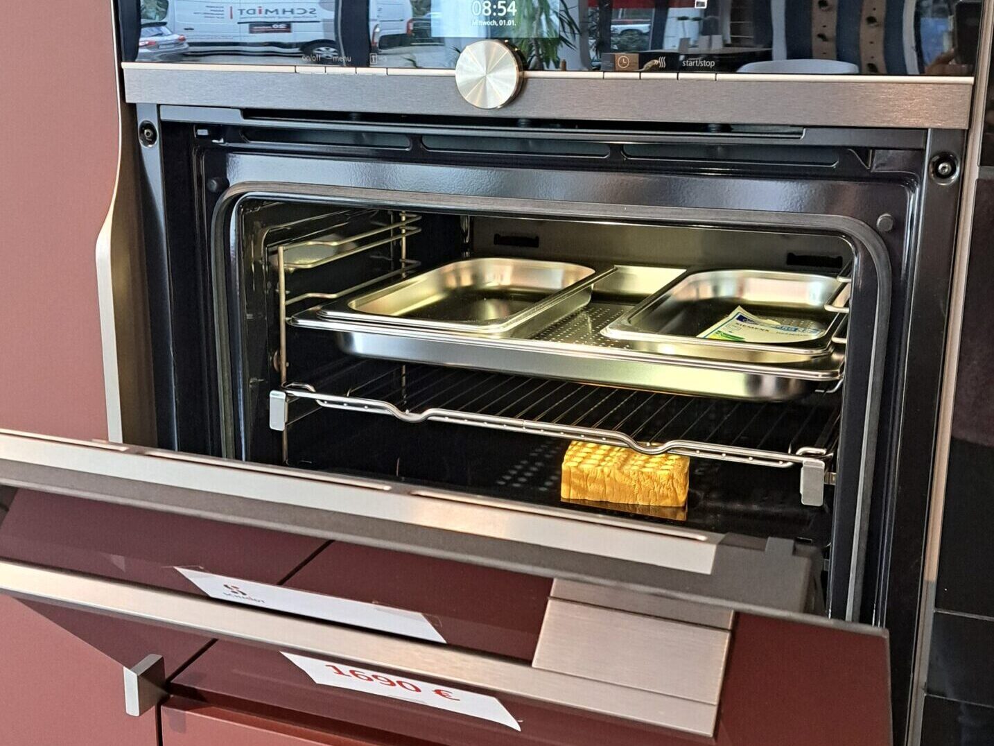 Ein Einbaubackofen mit teilweise geöffneter Tür, in der Backbleche und ein Laib Brot zu sehen sind. Der Backofen ist in einem kastanienbraunen Schrank mit modernem Design untergebracht.