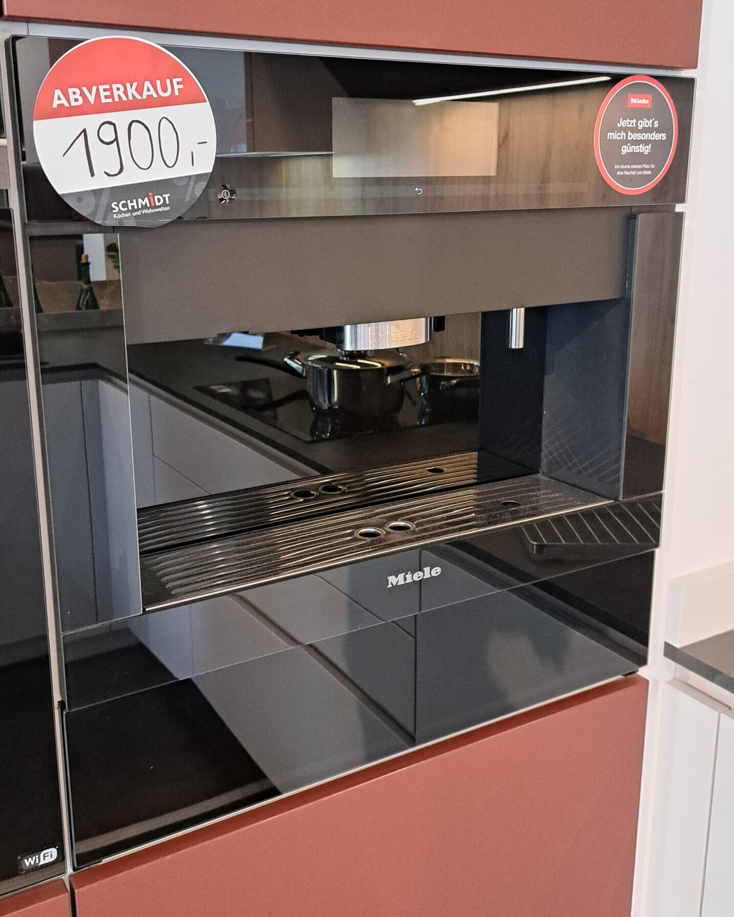 Ein schwarzer Kaffeeautomat als Einbaugerät von Miele mit spiegelnder Oberfläche steht in einem Ausstellungsraum. Auf einem roten Verkaufsschild steht ABVERKAUF 1900,-. Eine moderne Küche mit Herd und Töpfen spiegelt sich im Glas.