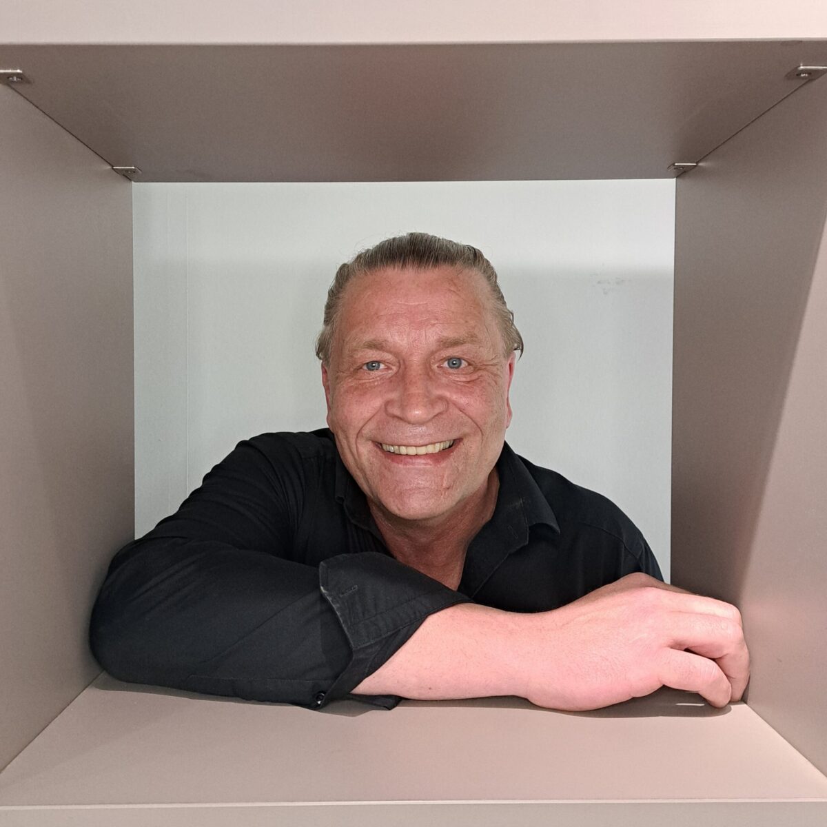 Ein lächelnder Mann mit zurückgekämmtem Haar und schwarzem Hemd beugt sich vor und blickt durch ein quadratisches, beigefarbenes Regal oder einen Rahmen. Der Hintergrund ist schlicht und in hellen Farben gehalten.