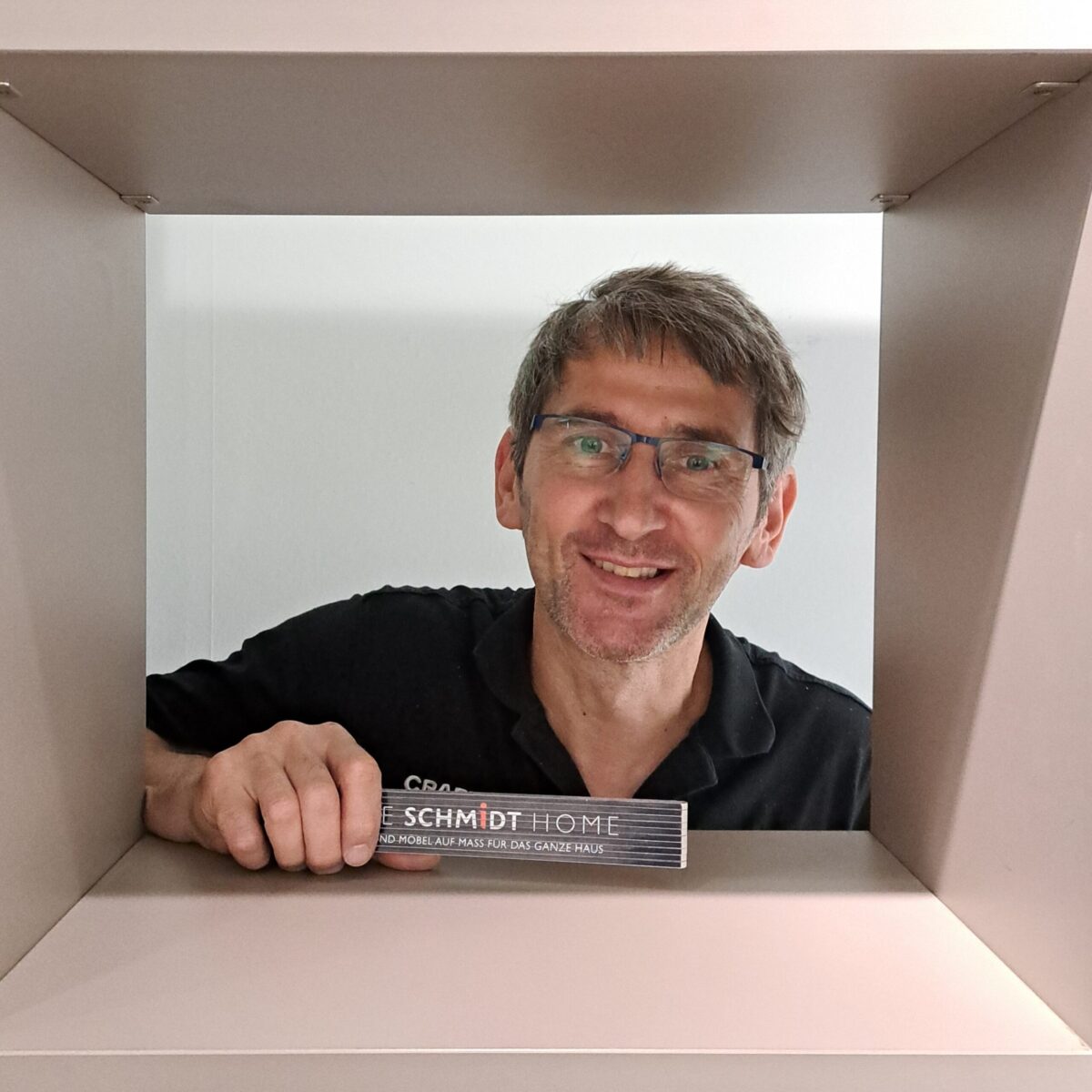 Ein Mann mit Brille und Bart lächelt, während er eine "Schmidt Home"-Broschüre in der Hand hält, umrahmt von einem quadratischen, beigen Bauwerk, als würde er durch ein Fenster schauen.