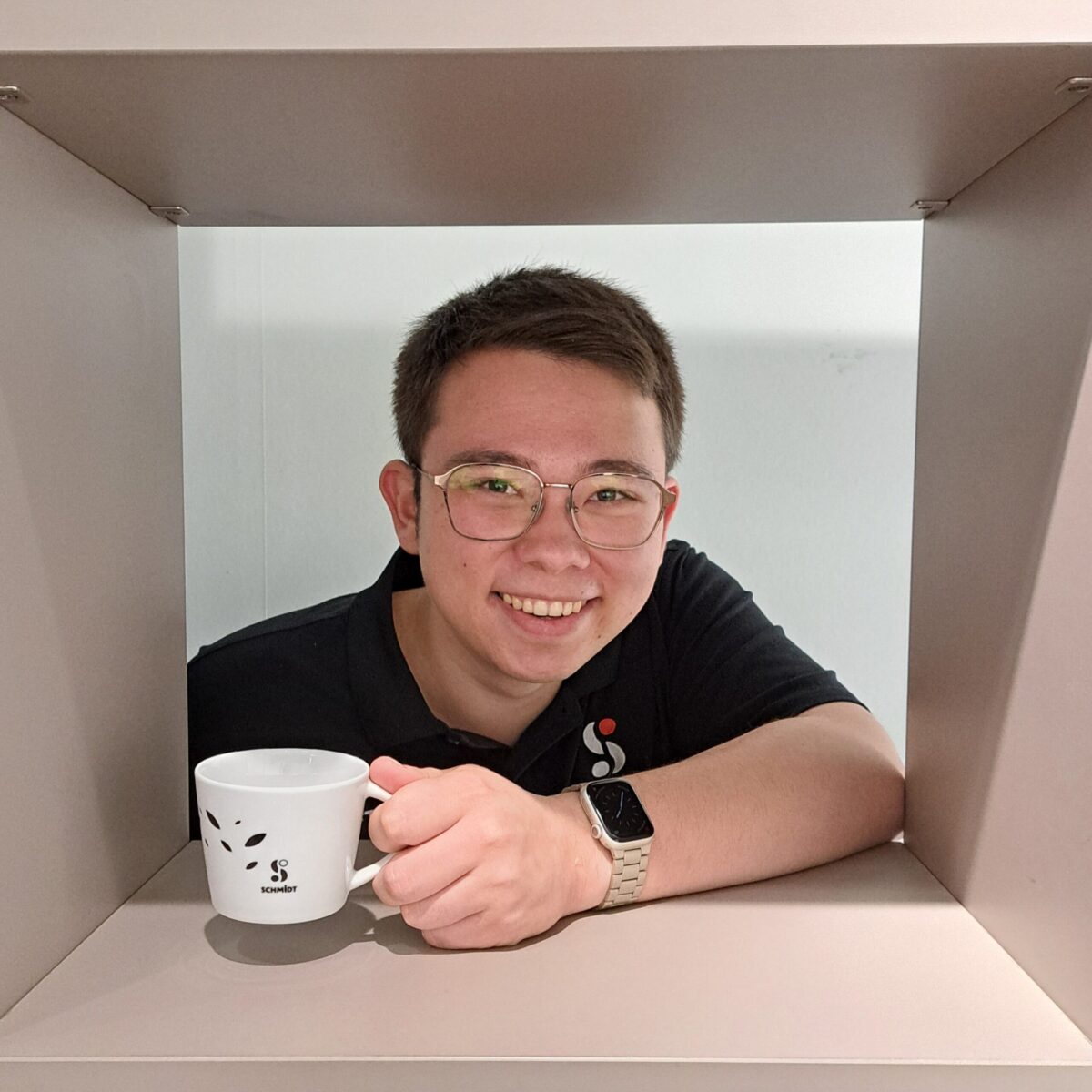 Eine Person mit kurzen Haaren und Brille lächelt, während sie einen weißen Becher hält. Sie trägt ein schwarzes Polohemd und eine Smartwatch und sitzt hinter einer quadratischen Regalöffnung mit einfarbigem Hintergrund.