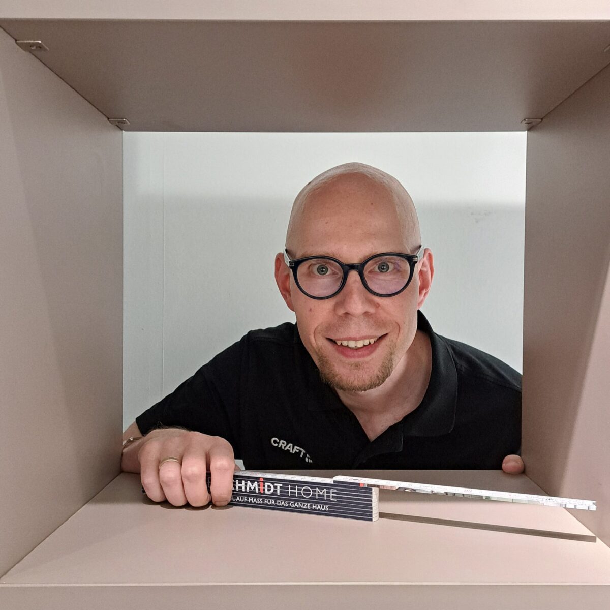 Ein lächelnder Mann mit Glatze und Brille blickt durch eine rechteckige Öffnung in einer beigen Struktur, in der Hand ein Buch und ein Messgerät. Er trägt ein schwarzes Hemd mit der Aufschrift CRAFT am Kragen.
