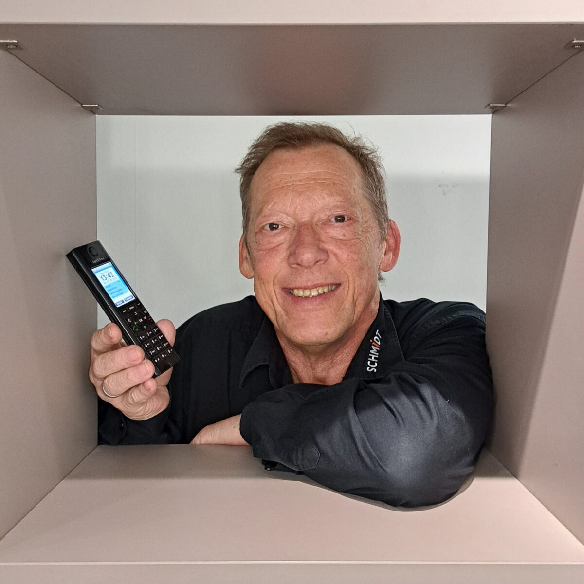 Ein lächelnder Mann in einem schwarzen Hemd lehnt an einem Regal, hält ein schnurloses Telefon hoch und blickt durch eine quadratische Öffnung direkt in die Kamera.
