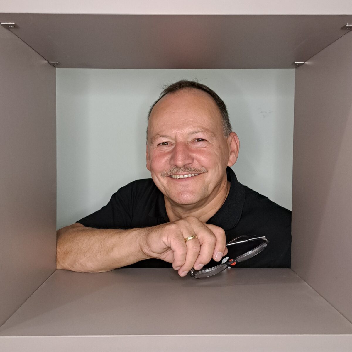 Ein lächelnder Mann mittleren Alters mit Schnurrbart, der ein schwarzes Hemd trägt, ist durch einen quadratischen grauen Rahmen zu sehen. Er hält eine Brille in einer Hand und stützt seine Arme auf den unteren Rand des Rahmens.
