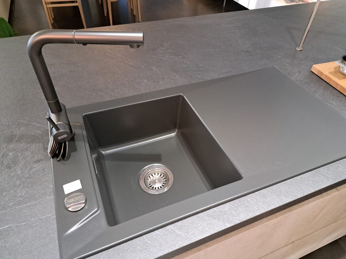 Ein modernes, mattschwarzes Spülbecken mit einem passenden Wasserhahn ist in eine dunkle Arbeitsplatte eingelassen. Neben dem Waschbecken befinden sich ein Seifenspender und ein Abflussdeckel. Der Hintergrund zeigt einen Teil einer Küche mit Holzmöbeln.