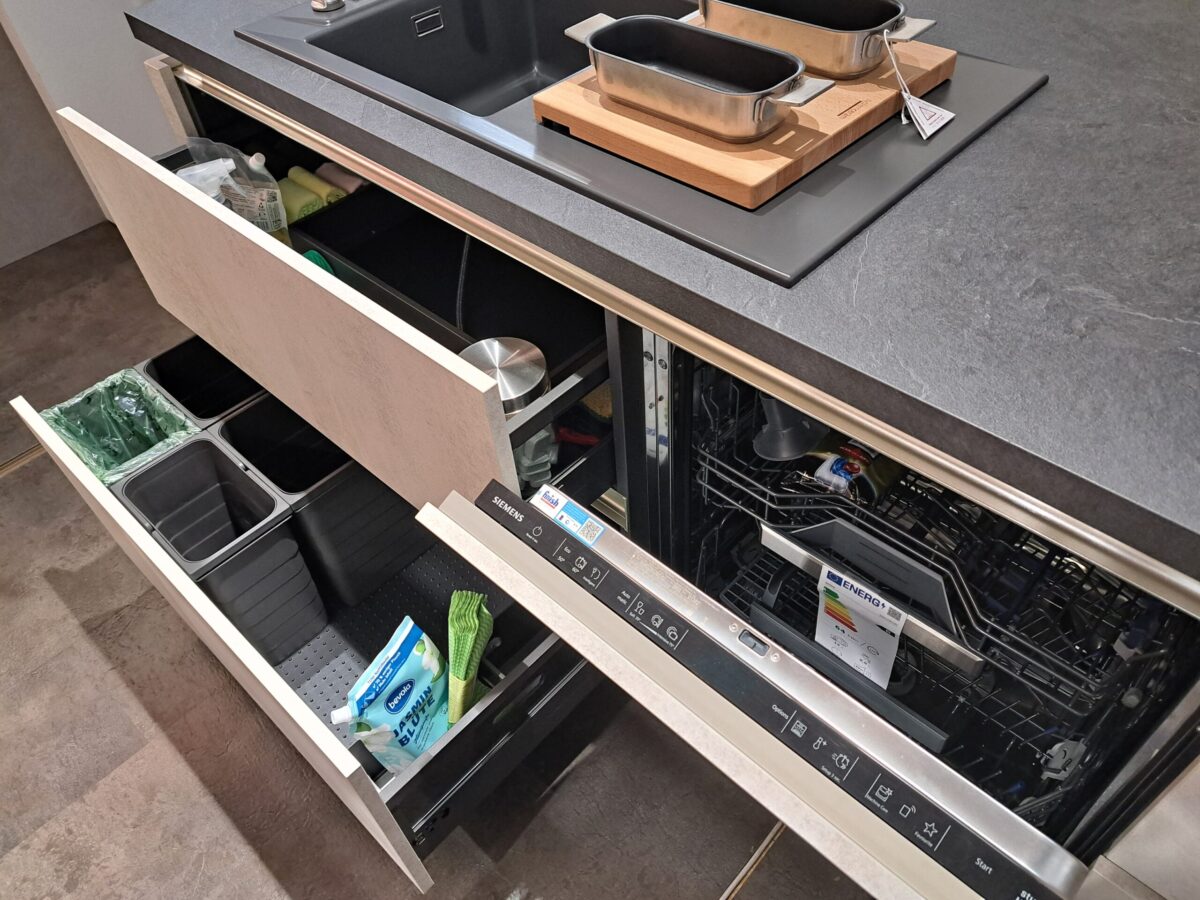 Eine moderne Küche mit offenen Schubladen, die geordnete Abfallbehälter, Reinigungsmittel und einen offenen eingebauten Geschirrspüler mit Geschirr unter einer Spüle und einem Herd mit zwei Pfannen auf einem Schneidebrett zeigen.