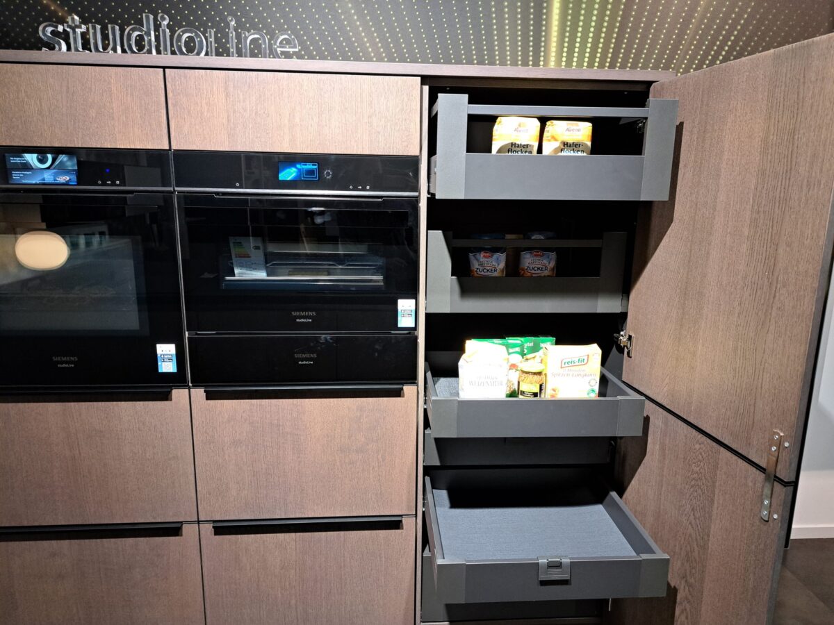 Eine moderne Küche mit dunklen Holzschränken, Einbaubacköfen auf der linken Seite und einem offenen Vorratsschrank auf der rechten Seite mit vier ausziehbaren Regalen mit verschiedenen Lebensmittelverpackungen.