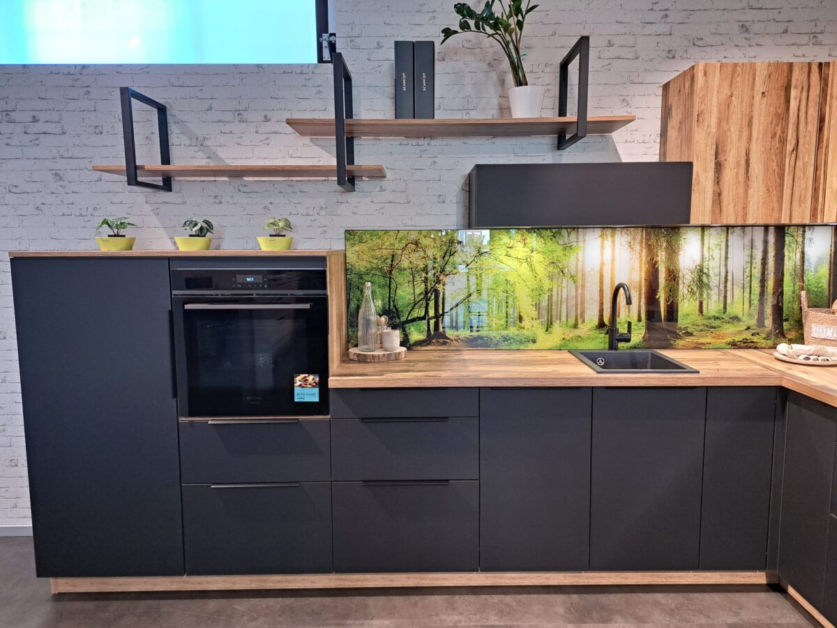Moderne Küche mit mattschwarzen Schränken, Holzakzenten, offenen Regalen mit Topfpflanzen und einer Nischenrückwand mit dem Motiv einer Waldszene. Ein eingebauter Ofen, eine Spüle und eine Arbeitsplatte sind sichtbar.
