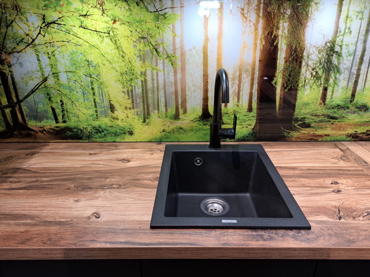 Eine moderne schwarze Küchenspüle mit einem hohen Wasserhahn ist in eine Arbeitsplatte mit Holzoptik eingelassen. Hinter der Spüle ist eine Nischenrückwand auf der eine lebhafte Waldszene mit sonnenbeschienenen grünen Bäumen zu sehen ist.