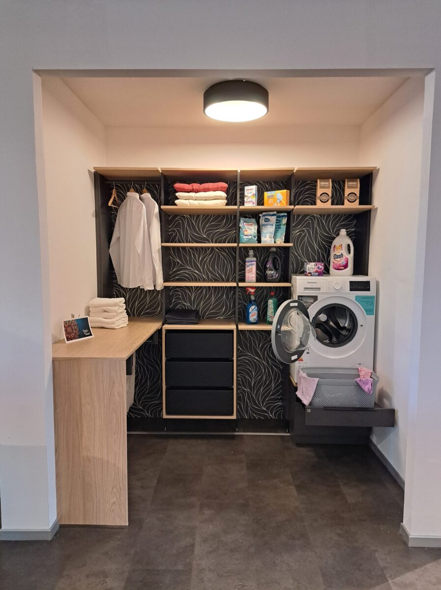 Eine moderne Waschküche mit offenen Regalen für Reinigungsmittel, gefaltete Handtücher und Kleidung auf Bügeln.