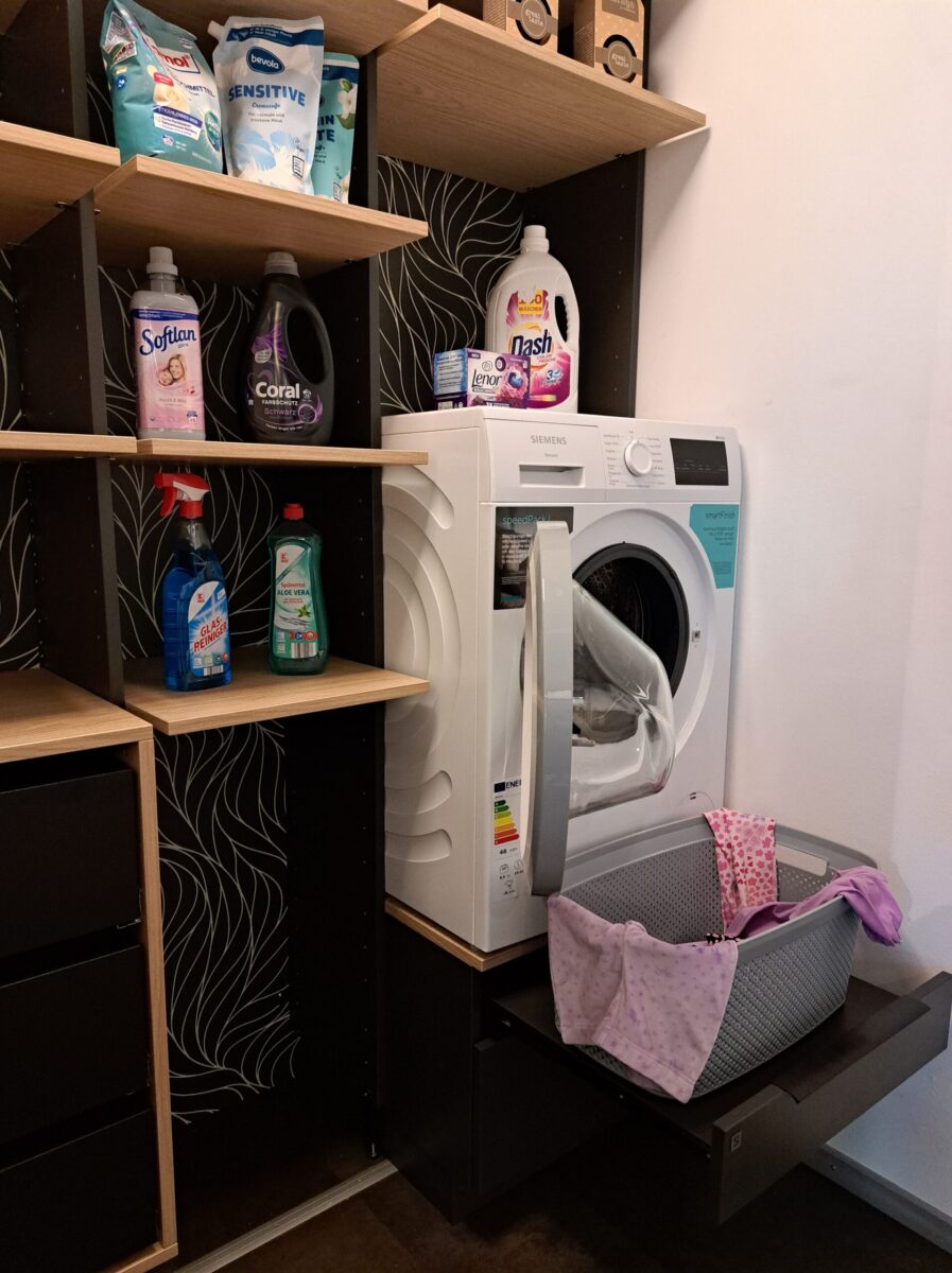 In der Ausstellung steht ein Hauswirtschaftsraum mit Waschküche. Die weiße Waschmaschine mit offener Tür steht erhöht und darunter befindet sich ein ausziehbares Tableau für den Wäschekorb. In den Regalen darüber sind Waschmittel, Weichspüler und Reinigungsmittel ordentlich aufbewahrt.
