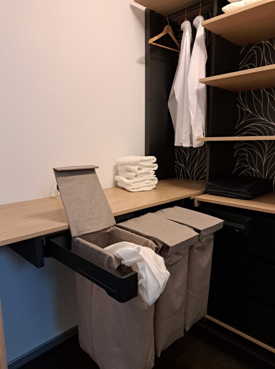 Ein moderner Kleiderschrank mit Holzregalen, gefalteten weißen Handtüchern, hängenden weißen Hemden und beigen Aufbewahrungen für Schmutzwäsche unter einer hölzernen Arbeitsplatte.