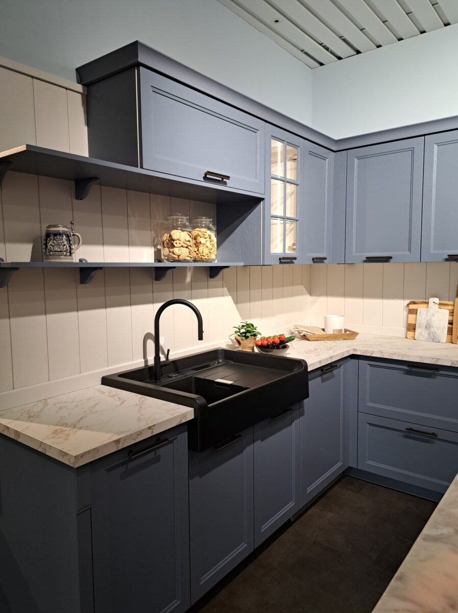 Eine moderne Ausstellungsküche im Landhaus-Stil mit hellblauen Schränken, Arbeitsplatten in Marmoroptik, einem schwarzen Spülbecken und offenen Regalen. Glasgefäße, eine Gemüseschale und Schneidebretter verleihen diesem einladenden Raum Charme.