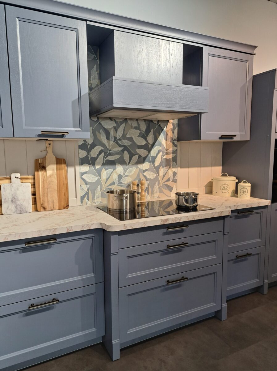 Moderne Landhausküche mit hellblauen Schränken, einer Küchenarbeitsplatte in Marmoroptik und einem Herd. Die Rückwand weist ein graues und weißes Blattmuster auf, während hölzerne Schneidebretter die Theken schmücken.