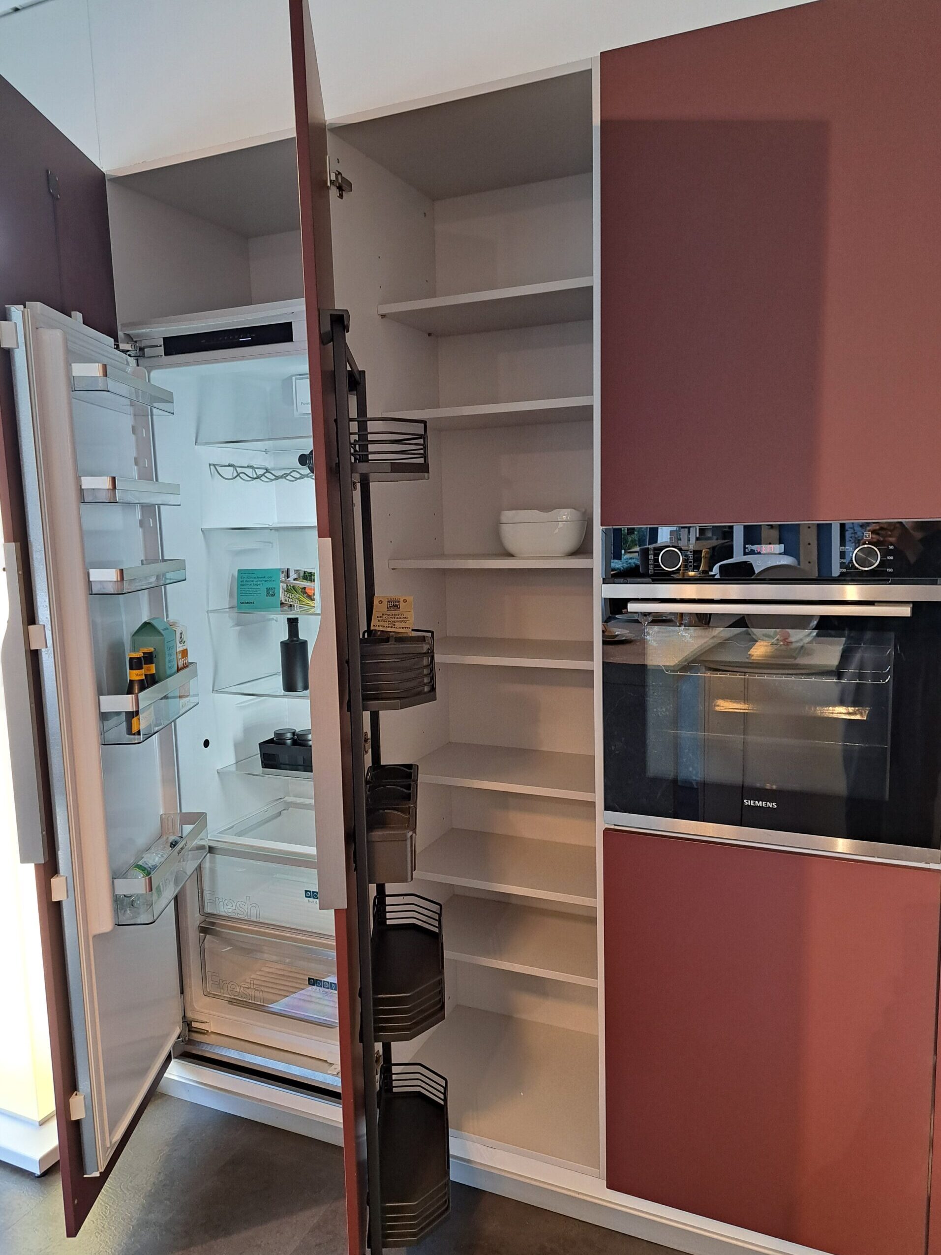 Ein moderner Küchenschrank mit offenen Türen gibt den Blick auf einen weitgehend leeren Kühlschrank, einen Schrank mit viel Stauraum und einen Backofen frei.