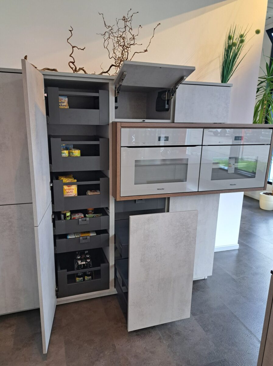 Moderne Abverkaufsküche mit grauen Schränken, von denen einige offen sind und den Blick auf ausziehbare Vorratsregale freigeben, in denen Lebensmittel aufbewahrt werden können. Die Einbaubacköfen sind sichtbar.
