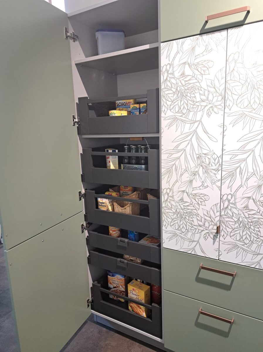 Ein geöffneter Küchenhochschrank gibt den Blick frei auf fünf ausziehbare Regalböden, die mit Lebensmitteln, Gläsern und Schachteln bestückt sind. Der Schrank daneben besticht durch ein auffälliges Printmuster.