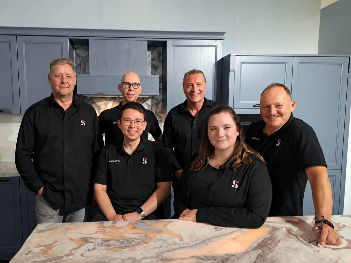 Sechs Teammitglieder in einheitlichen schwarzen Shirts mit dem Logo von „SCHMIDT Küchenstudio und Wohnwelten“ posieren lächelnd um eine Kücheninsel aus Naturstein in einer Landhausküche mit eleganten blauen Schränken.